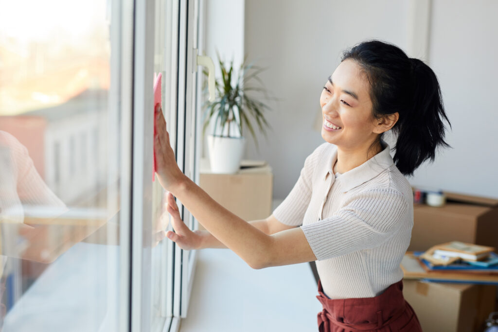 Waist up portrait of young Asian woman washing windows while enjoying Spring cleaning in house or apartment